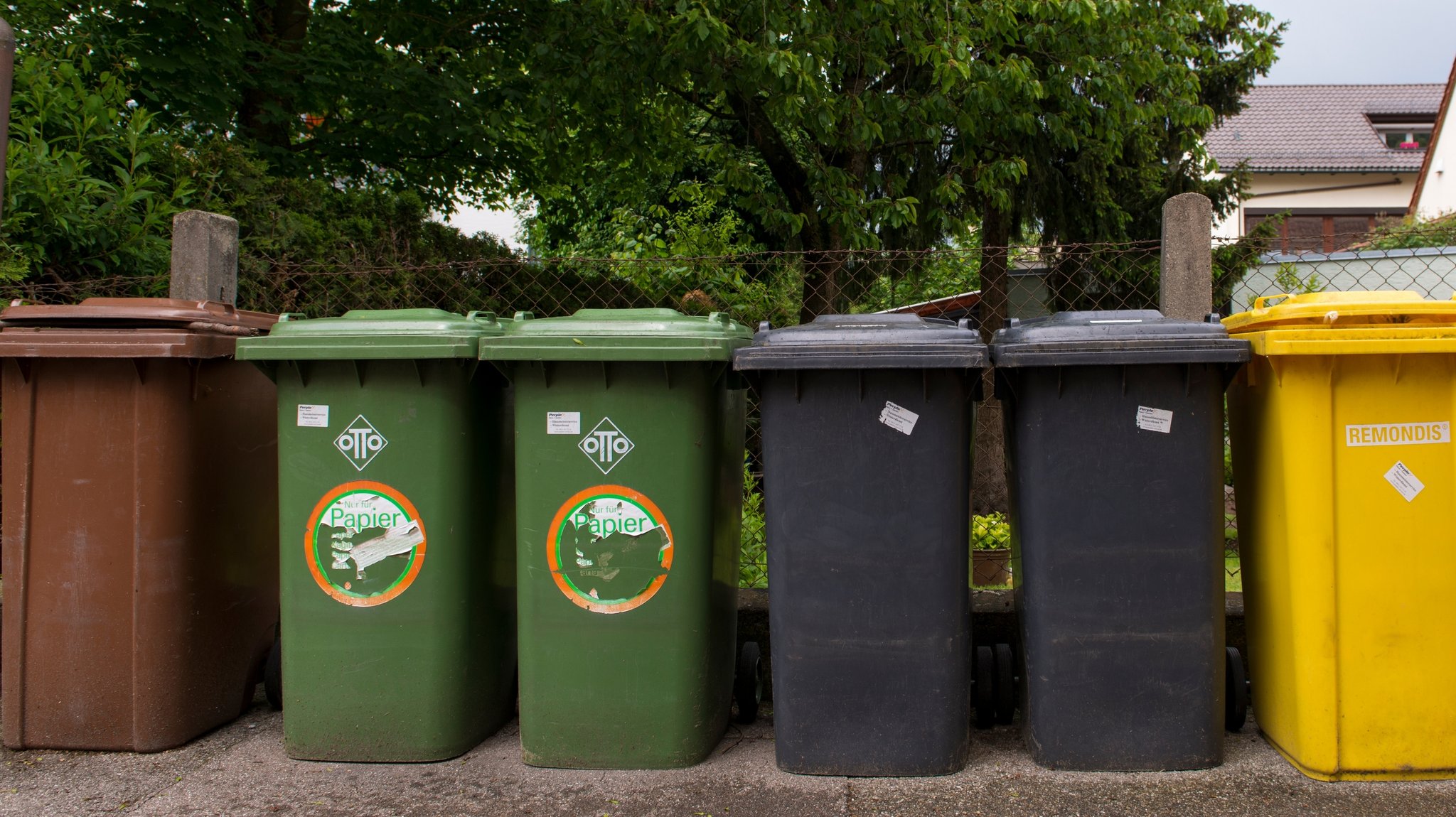 Mehrere Mülltonnen für Kompost, Papier, Restmüll und Verrkaufsverpackungen stehen nebeneinander an einem Zaun in Augsburg