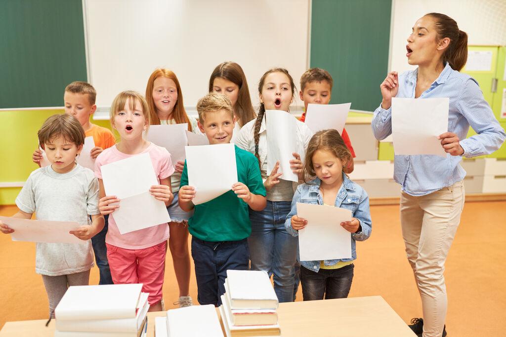 (Symbolbild) Grundschulkinder singen gemeinsam mit ihrer Lehrerin im Chor in einem Klassenzimmer.