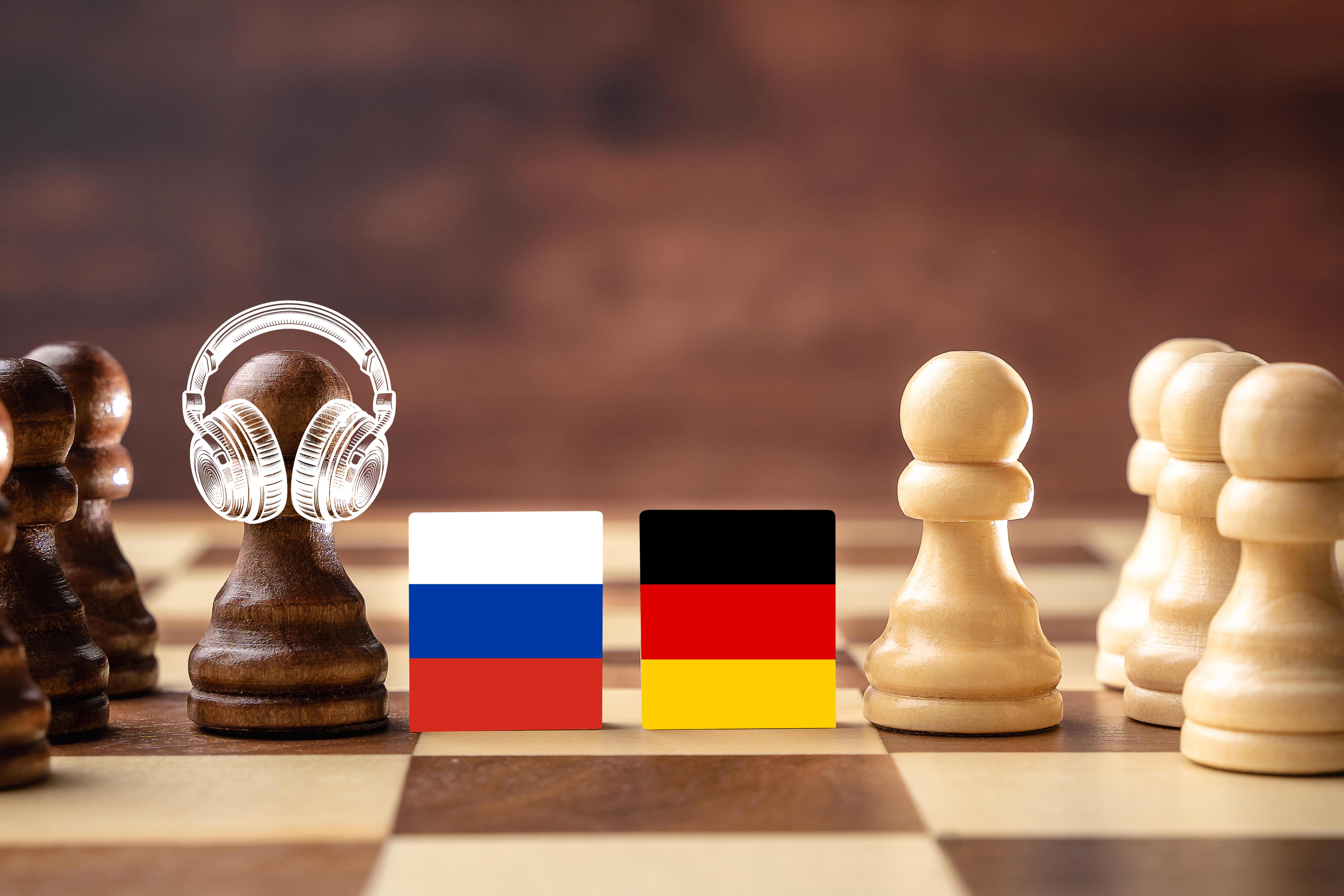 Bildmontage eines Schachbretts mit weißen und schwarzen Figuren, getrennt durch die russische und deutsche Flagge. Eine Figur auf der Seite der russischen Flagge trägt Kopfhörer.