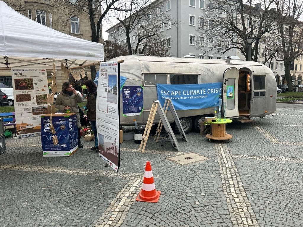 Ein Wohnwagen steht in Bayreuth herum. "Escape Climate Crisis" steht auf einem Banner, das am Wagen hängt. Daneben sind Roll-ups aufgestellt und zwei Frauen unterhalten sich. 
