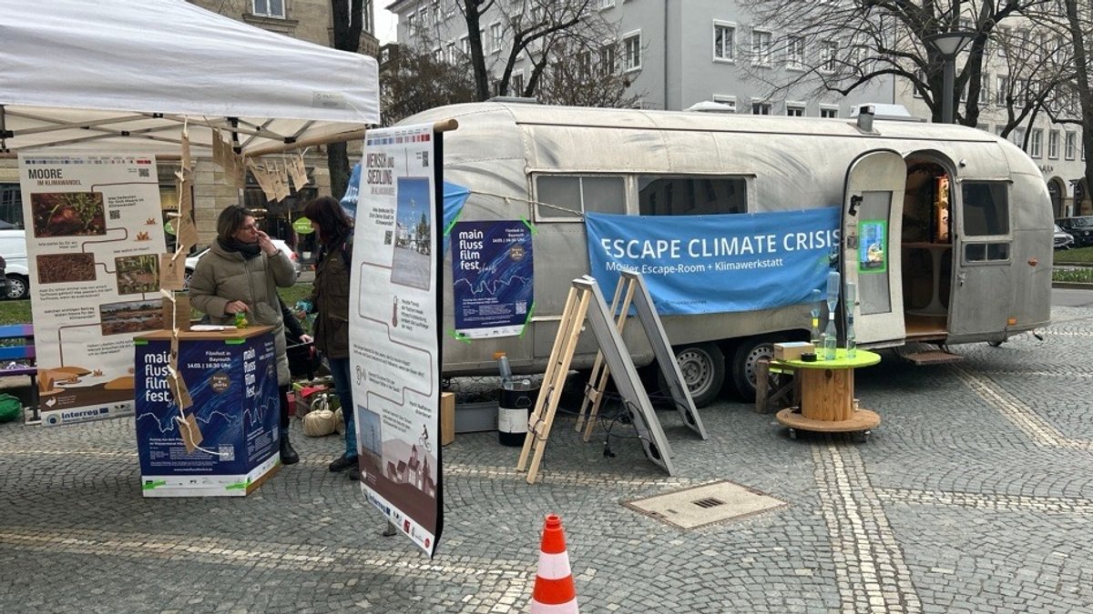 Ein Wohnwagen steht in Bayreuth herum. "Escape Climate Crisis" steht auf einem Banner, das am Wagen hängt. Daneben sind Roll-ups aufgestellt und zwei Frauen unterhalten sich. 