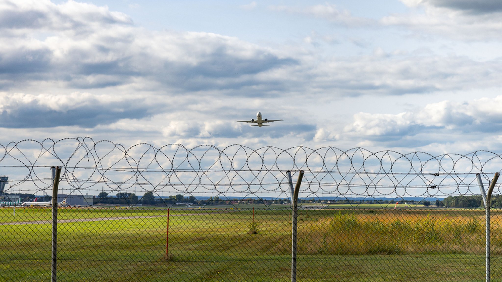 Sicherheitszaun am Flughafen Nürnberg mit Stacheldraht und einem Flugzeug, das gerade abhebt. Im Hintergrund sind Teile des Flughafens und der Kontrollturm zu sehen.