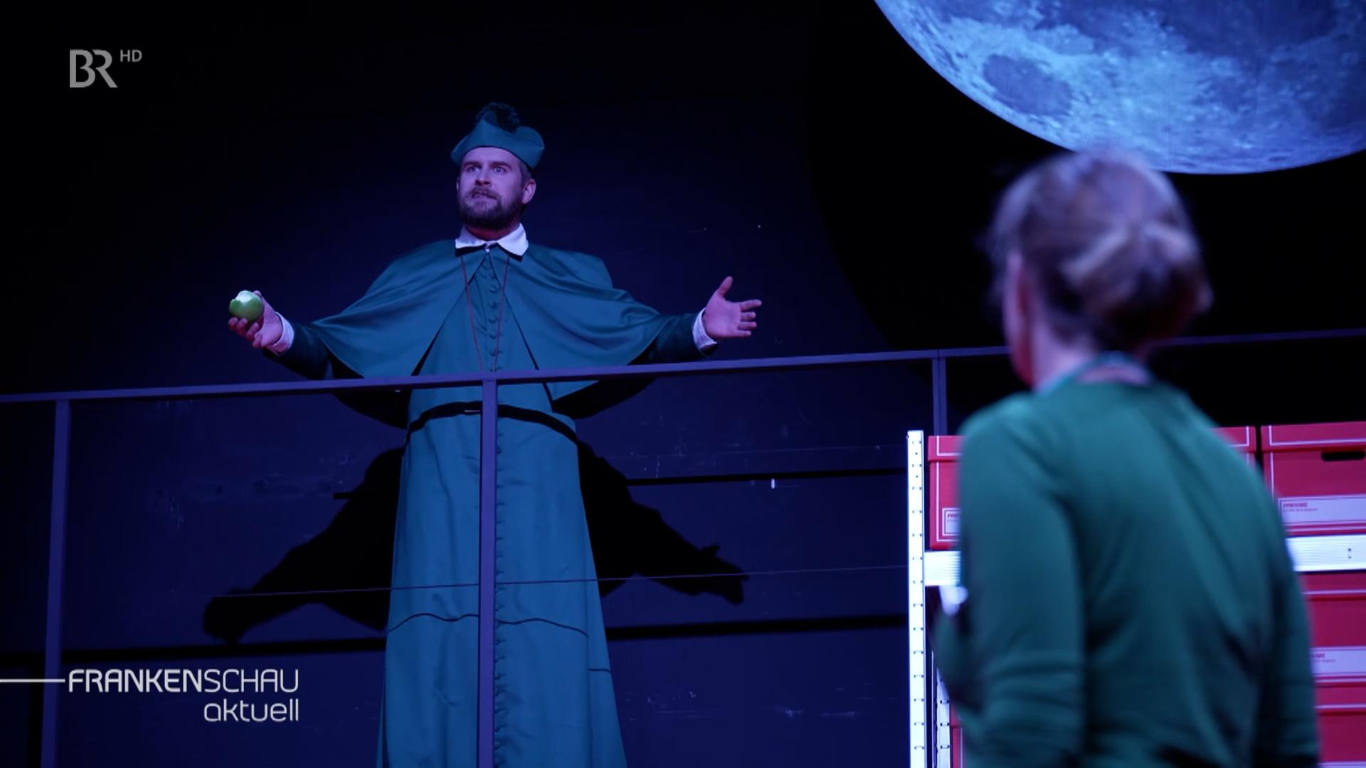 Ein Schauspieler im Talar eines Priesters, steht auf der Bühne und breitet die Arme aus. Vor ihm steht eine Frau im grünen Kleid.