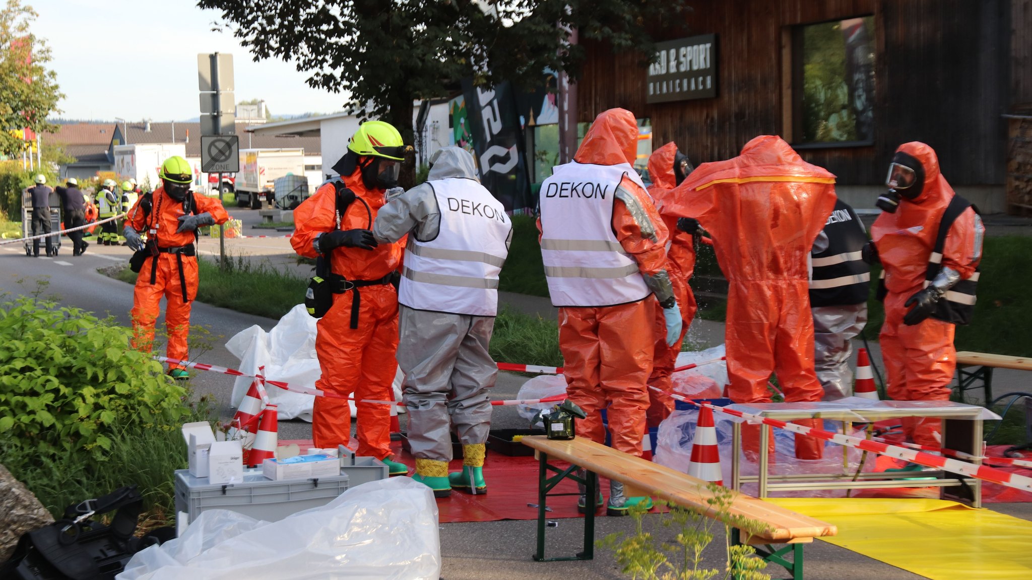 Feuerwehrkräfte stehen in orangen Spezialanzügen und mit Atemmasken nach dem Chemieunfall in Blaichach auf der Straße.