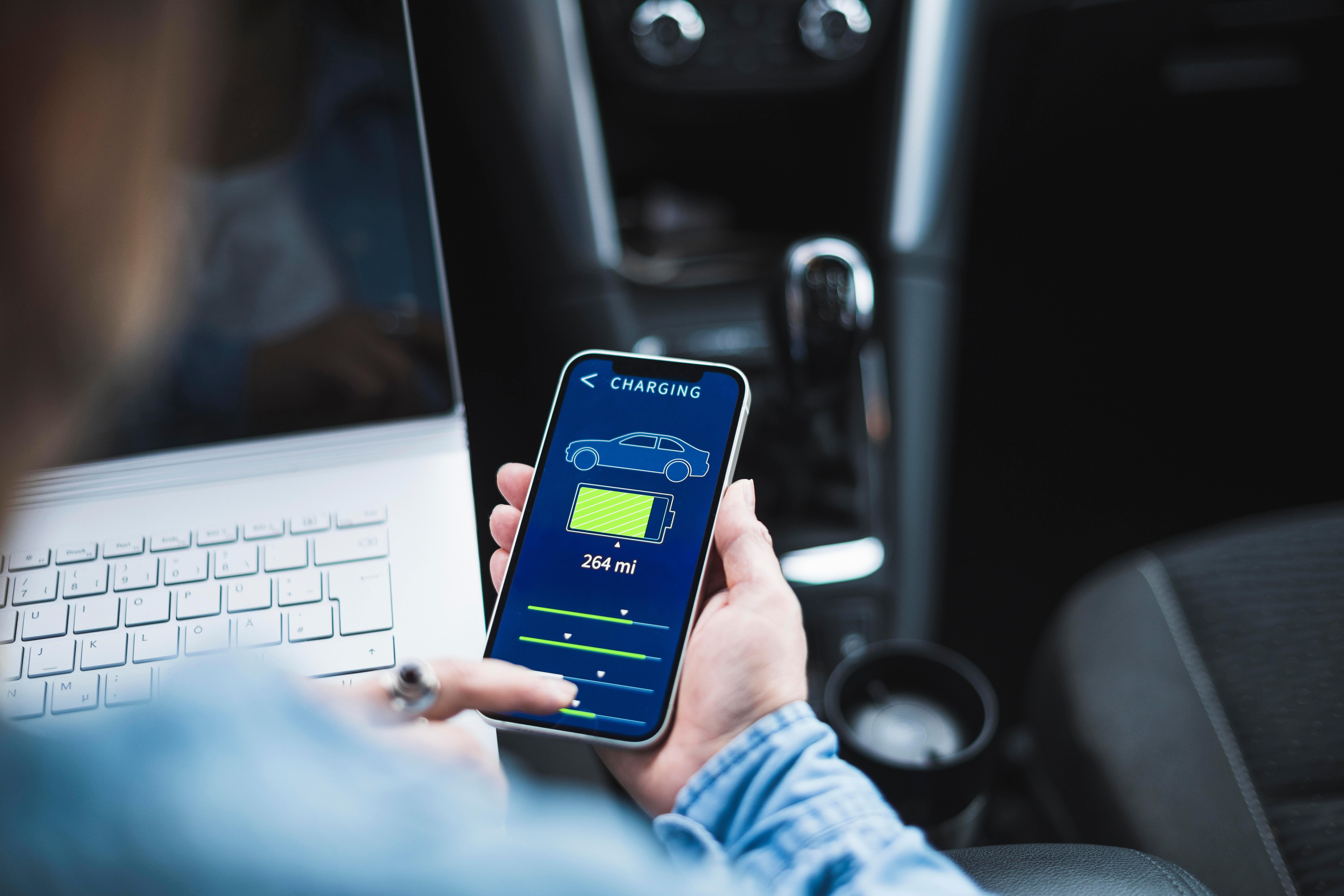 Eine Frau sitzt am Steuer ihres E-Autos mit dem Laptop auf den Knien und bedient an ihrem Smartphone die App, die das Laden der Autobatterie anzeigt.
