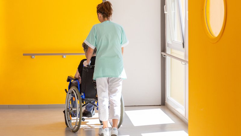 (Symbolbild) Eine Altenpflegerin schiebt eine im Rollstuhl sitzende Person. | Bild: stock.adobe.com/Reddragonfly (Symbolbild) Eine Altenpflegerin schiebt eine im Rollstuhl sitzende Person.