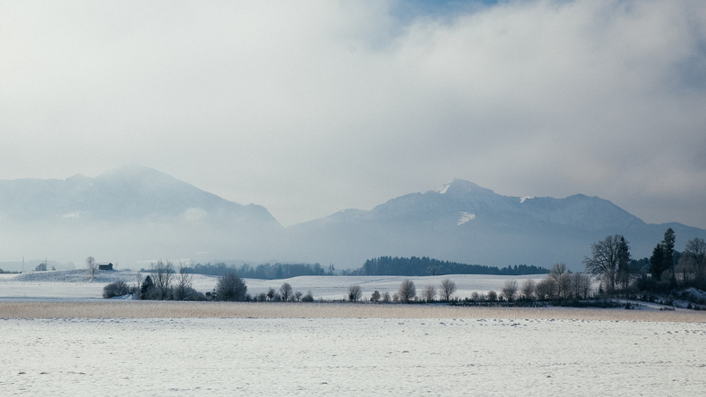 Chiemgauer Landschaft mit Blick auf verschneite Felder und Alpen in Wolken im Winter in Bayern.