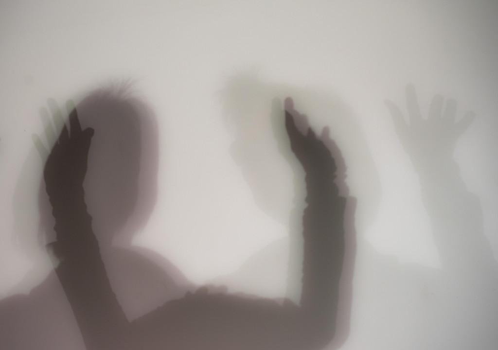 Symbolbild: Silhouette von zwei Menschen mit hochgestreckten Händen hinter einer trüben Scheibe.