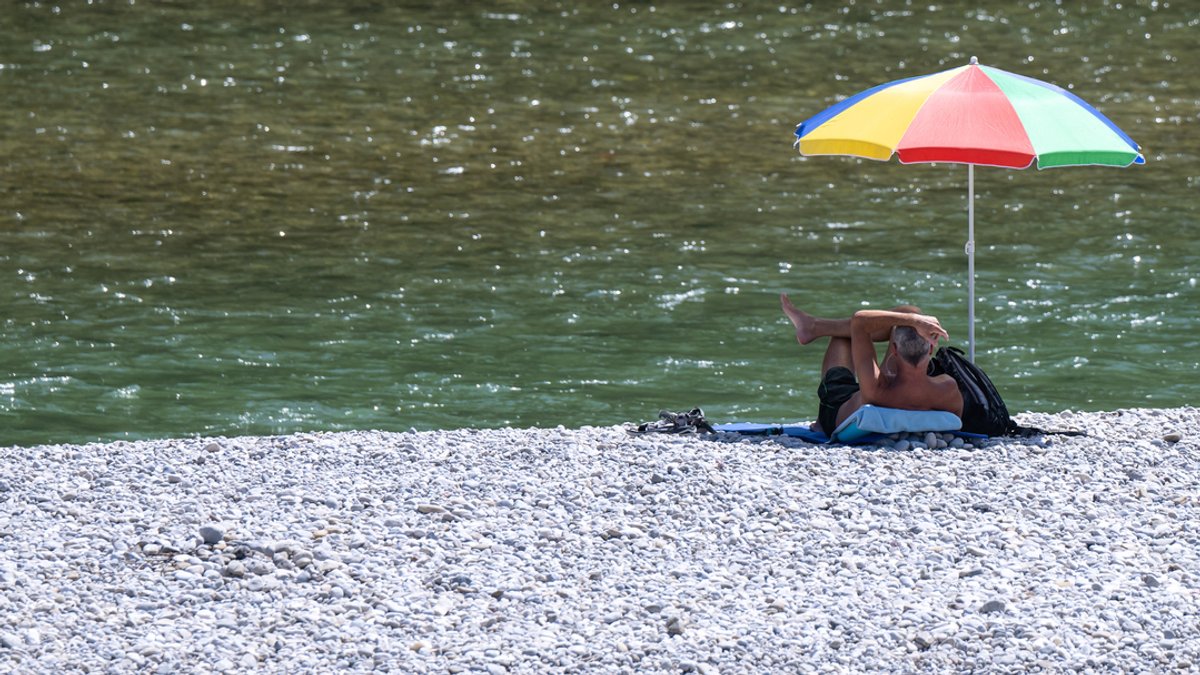 Ein Mann liegt unter einem bunten Sonnenschirm am Ufer der Isar in München.