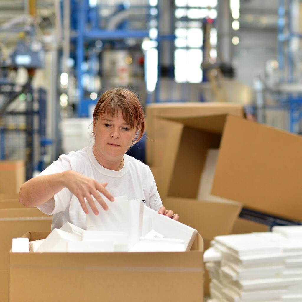 Symboldbild: Eine Frau steht in einer großen Lagerhalle und sortiert Ware aus einem großen Karton.