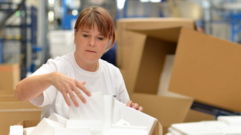 Symboldbild: Eine Frau steht in einer großen Lagerhalle und sortiert Ware aus einem großen Karton. | Bild: stock.adobe.com/industrieblick Symboldbild: Eine Frau steht in einer großen Lagerhalle und sortiert Ware aus einem großen Karton.