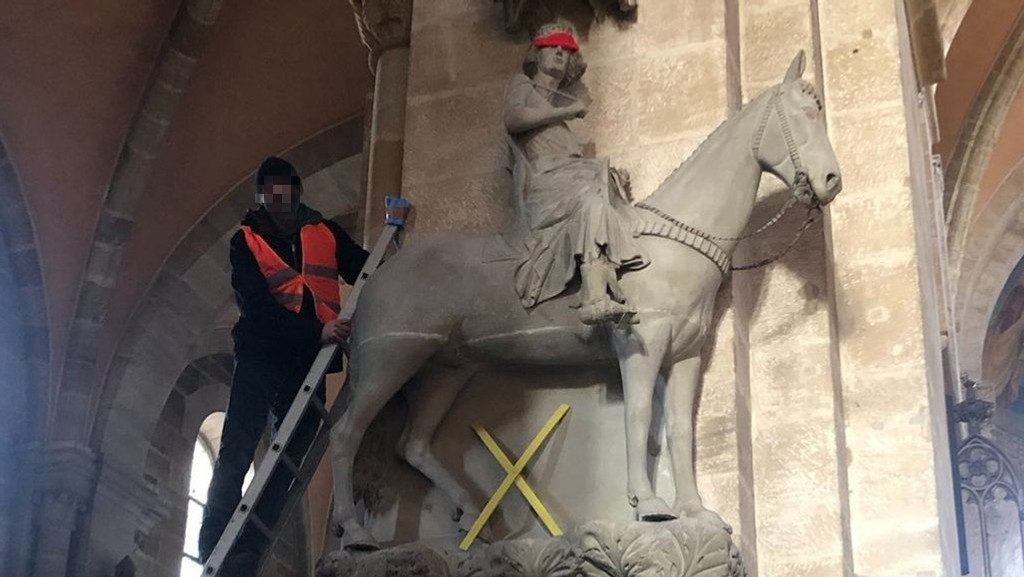 Ein Mann steht auf einer Leiter neben dem Bamberger Reiter im Bamberger Dom. Die Figur hat eine rote Augenbinde umgebunden bekommen.