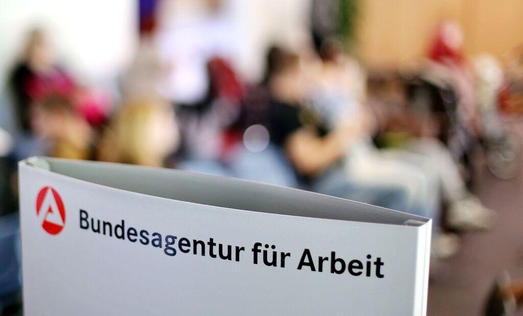 Ein Schild mit der Aufschrift "Bundesagentur für Arbeit" steht einem vollen Wartezimmer im Jobcenter.