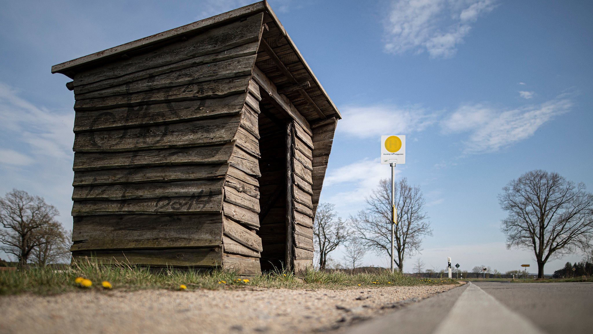 Eine Holzhütte steht als Bushaltehäuschen an einer Landstrasse.