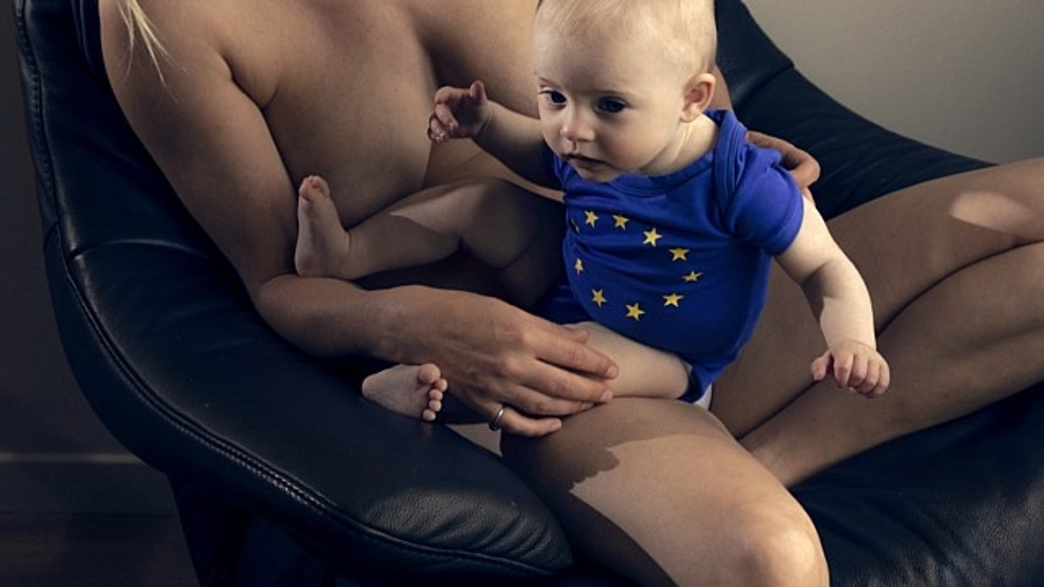 "EU Baby Jumper": Ein Baby im blauen Body mit gelben EU-Sternen sitzt auf dem Schoß einer nackten Frau.