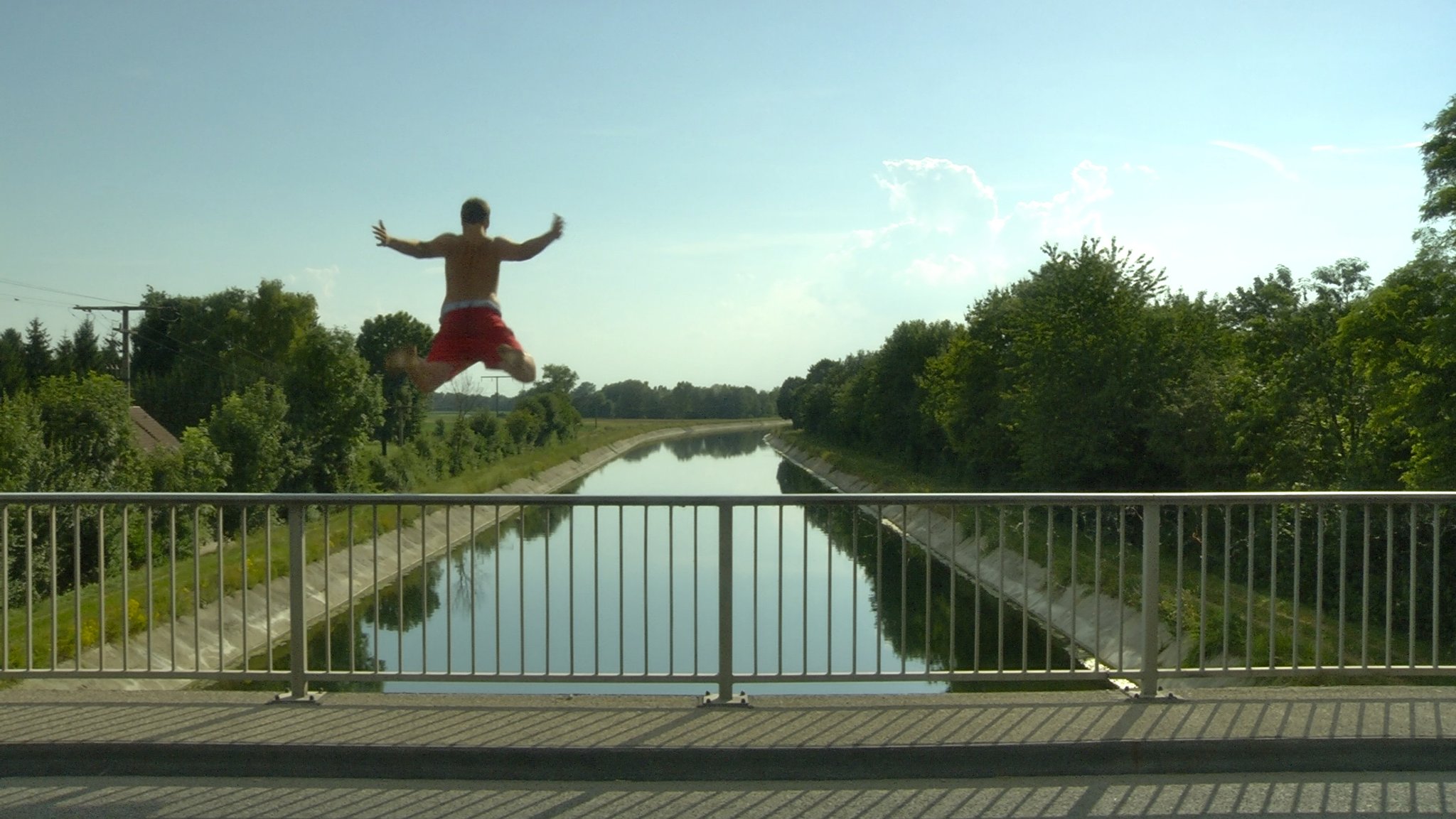 Ein junger Mann mit roter Badehose springt im Sommer von einer Brücke in einen Fluss