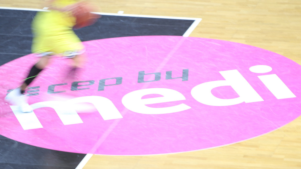 Ein Medilogo klebt auf dem Parkett eines Basketballfelds.
