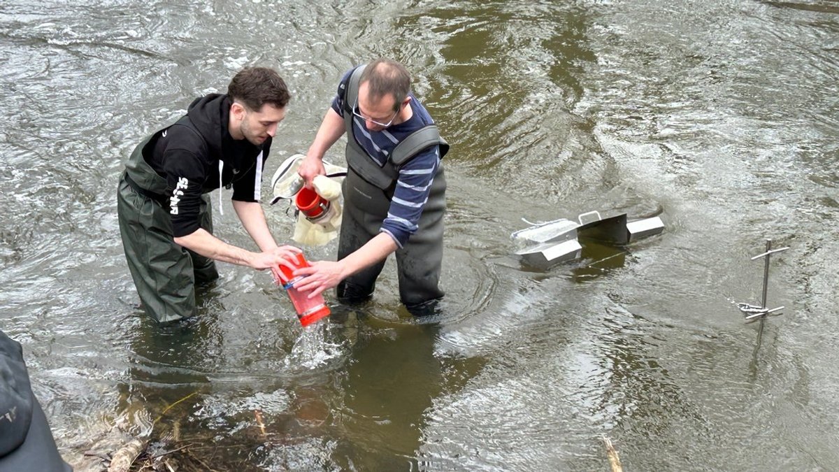 Zwei Männer stehen in Watthosen in einem Fluss und filtern Wasser durch verschiedene Geräte. 