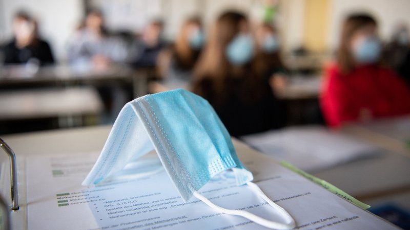 Eine Maske liegt im Unterricht auf Unterlagen, während im Hintergrund Schülerinnen und Schüler in einem Gymnasium mit Mund- und Nasenschutz zu sehen sind. 