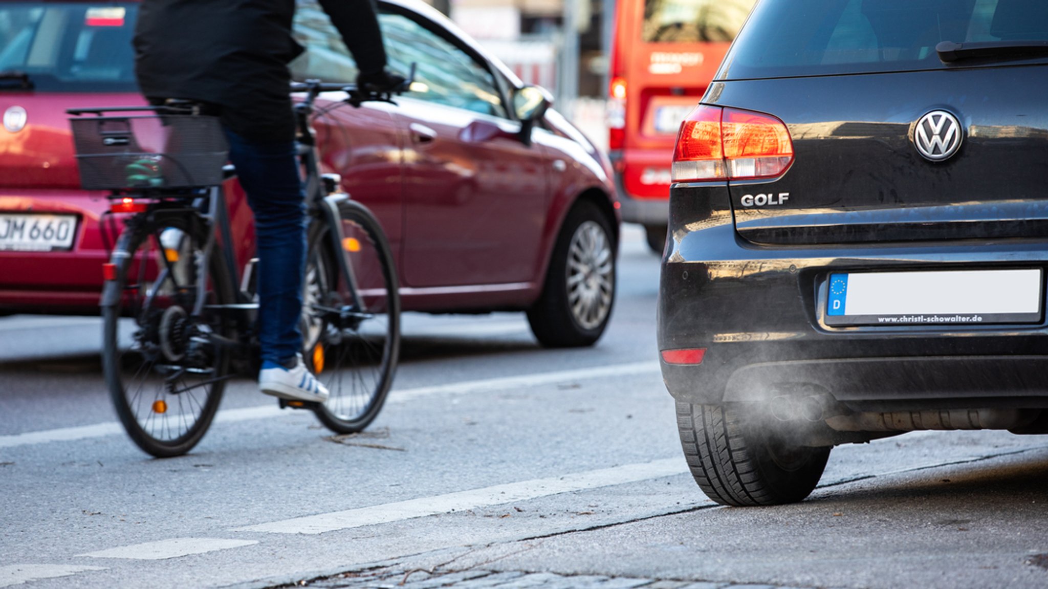 Ein Fahrradfahrer fährt an einem Auto vorbei, aus dessen Auspuff Abgase kommen.