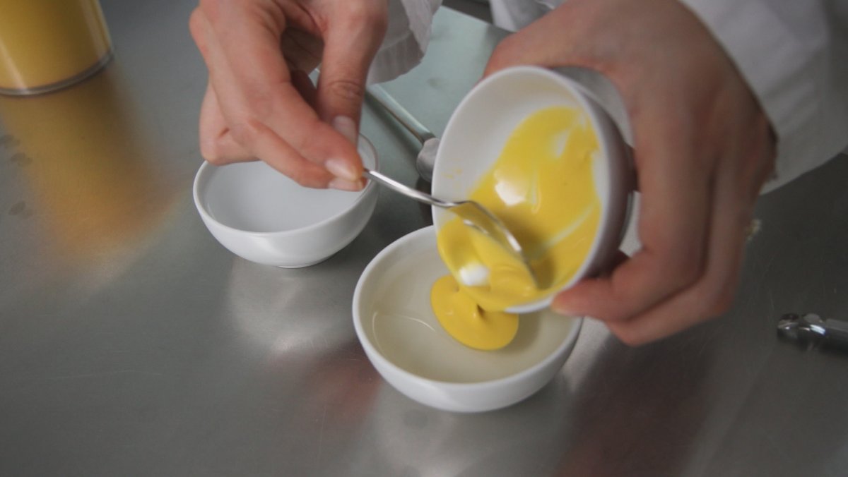 Eine Person schüttet flüssiges Eigelb aus einer Tasse in eine andere Tasse mit einer klaren Flüssigkeit. Das Eigelb bildet einen Kreis