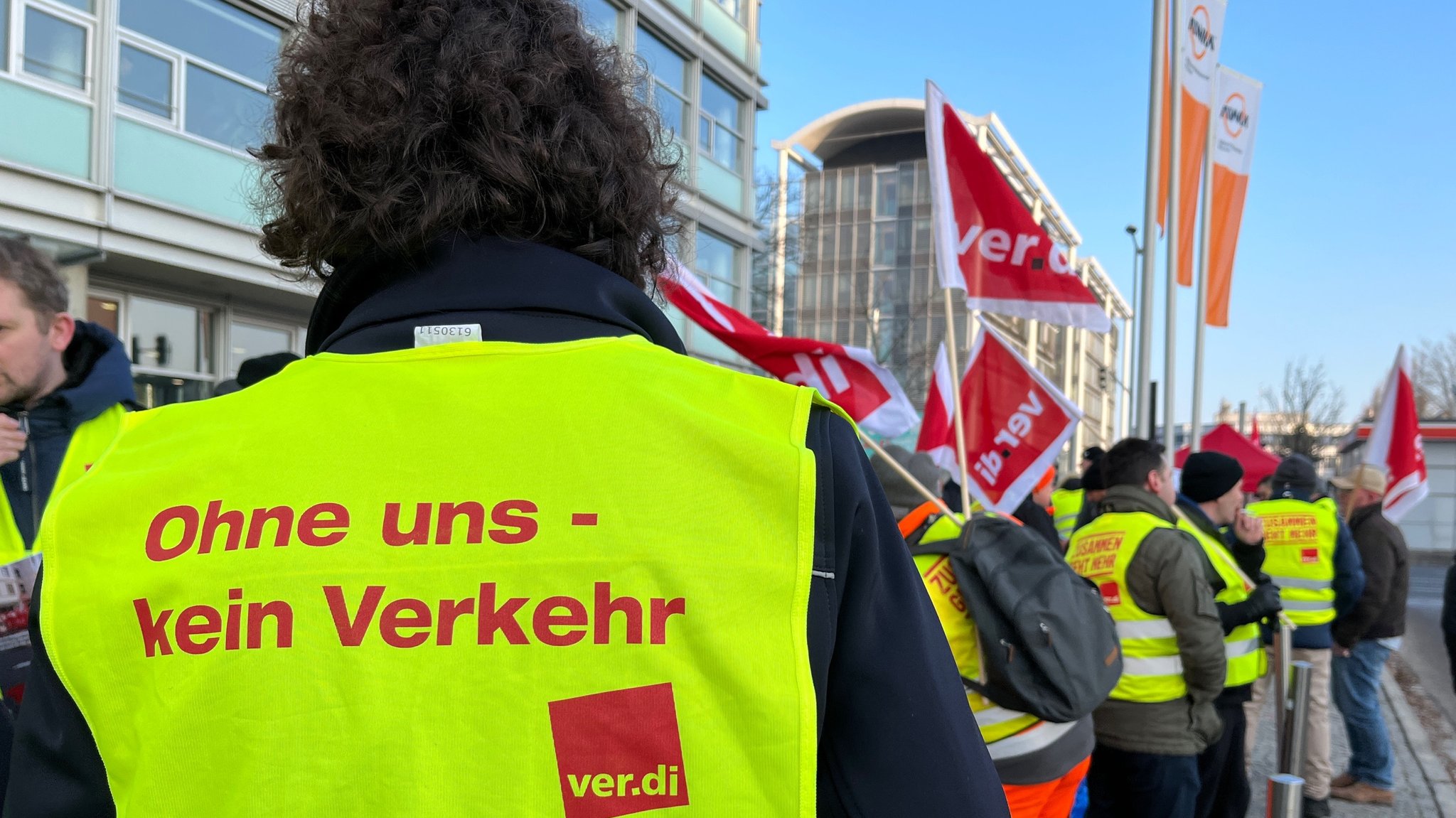 Beschäftigte in Warnwesten mit Verdi-Fahnen; im Vordergrund der Rücken einer Frau. Auf ihrer Warnweste ist zu lesen: "Ohne uns - kein Verkehr". Im Hintergrund in Unschärfe die Gebäude und Fahnen der Abfallwirtschaft München.