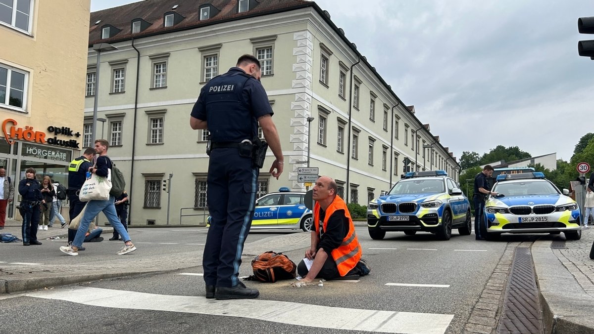 Ein Polizist steht am Montagnachmittag in Passau vor einem Mann, der sich auf dem Asphalt festgeklebt hat