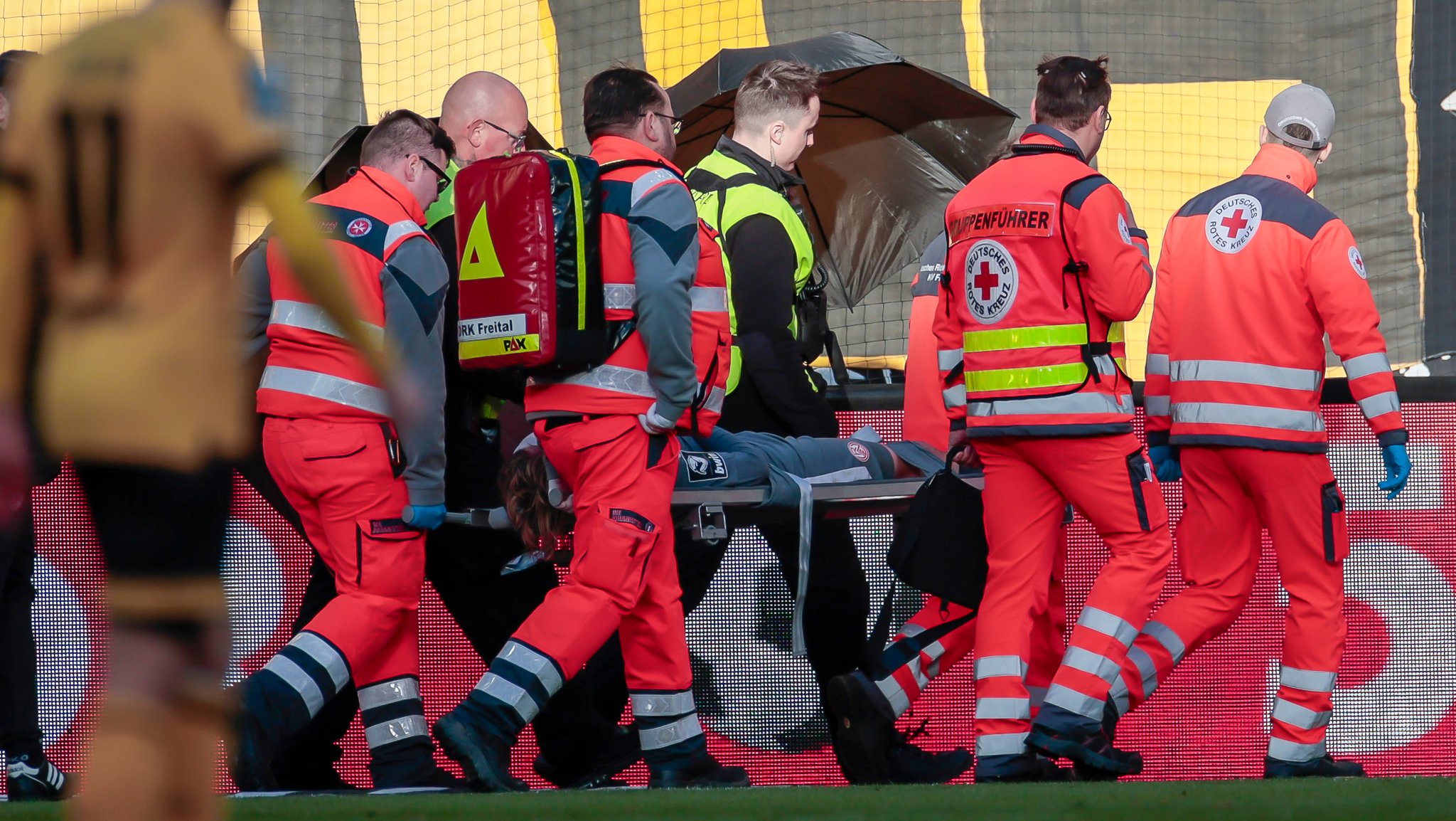 Felix Götze wird verletzt abtransportiert. Ordner müssen ihn mit Regenschirmen vor Wurfgeschossen schützen.