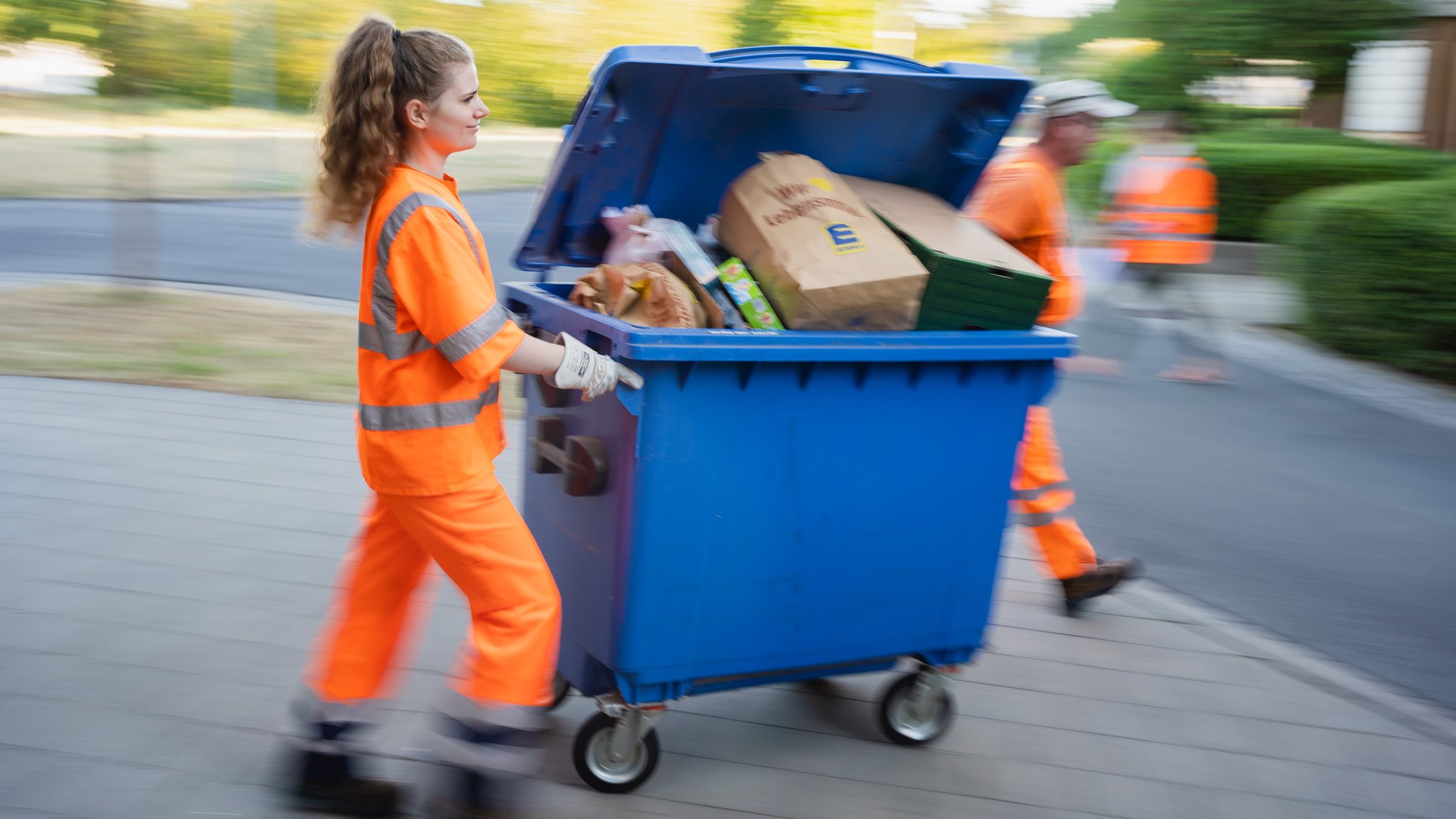 Eine Studentin in Warnkleidung schiebt eine Mülltonne.