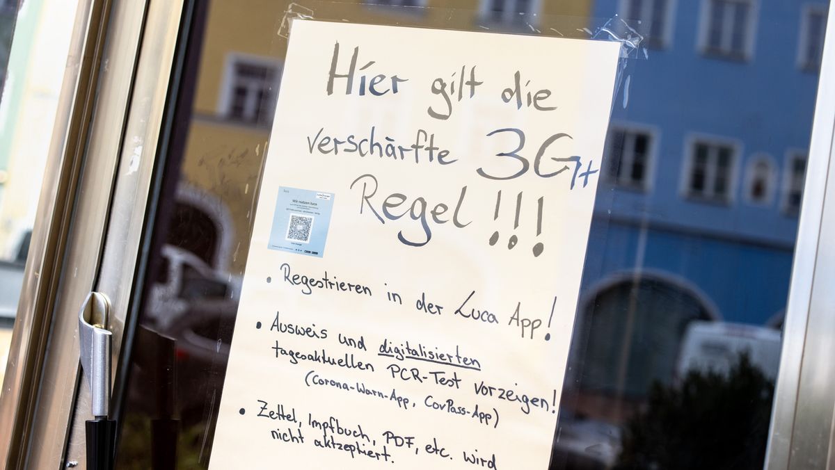 Auf einem Zettel an einer Scheibe von einem Café wird auf die Anwendung der verschärften "3G+ Regeln" hingewiesen, während sich in der Scheibe die für Mühldorf typischen Häuser im Inn-Salzach-Stil spiegeln.