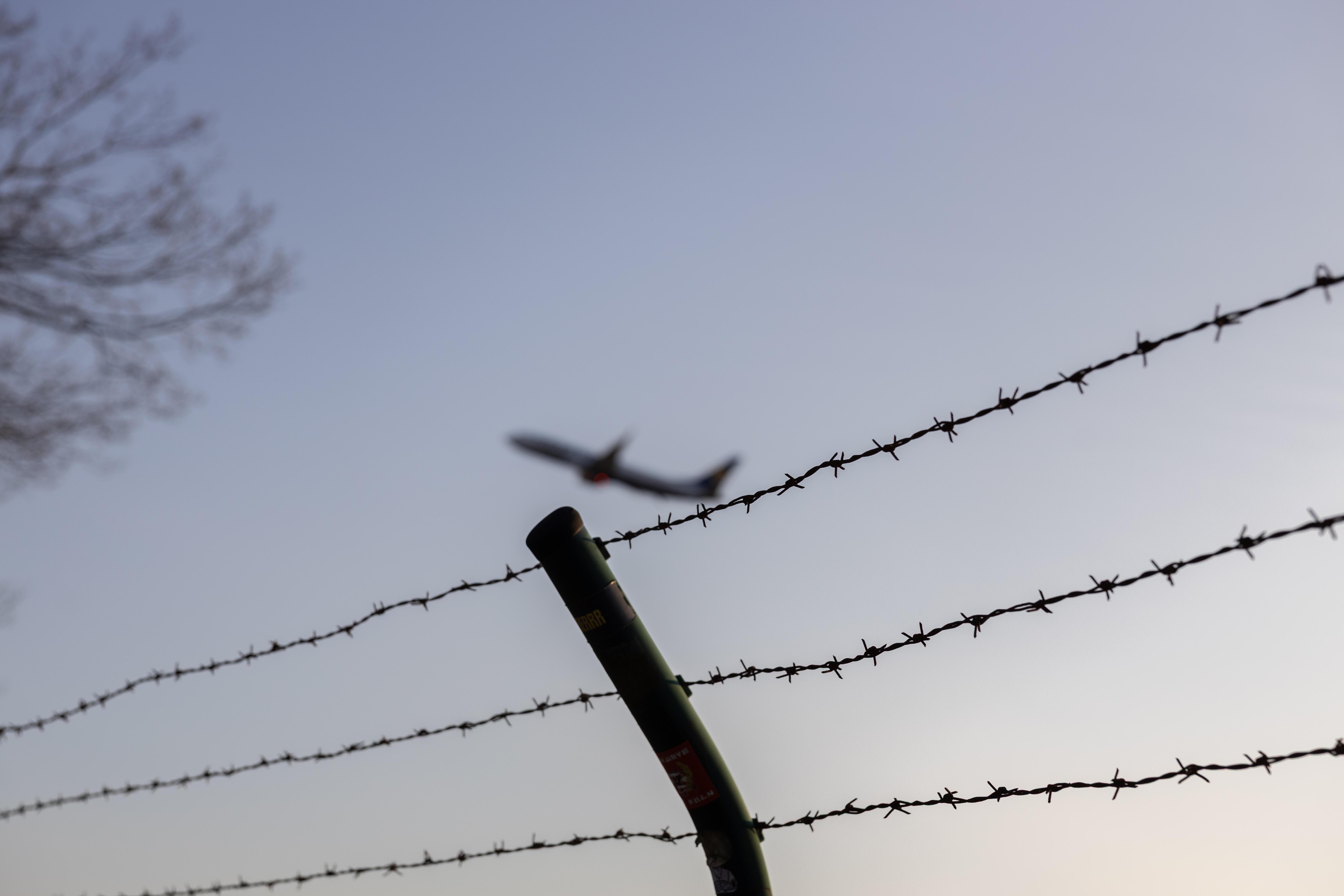 Ein Flugzeug fliegt in der Luft. Vordergründig sieht man einen Stacheldrahtzaun. 