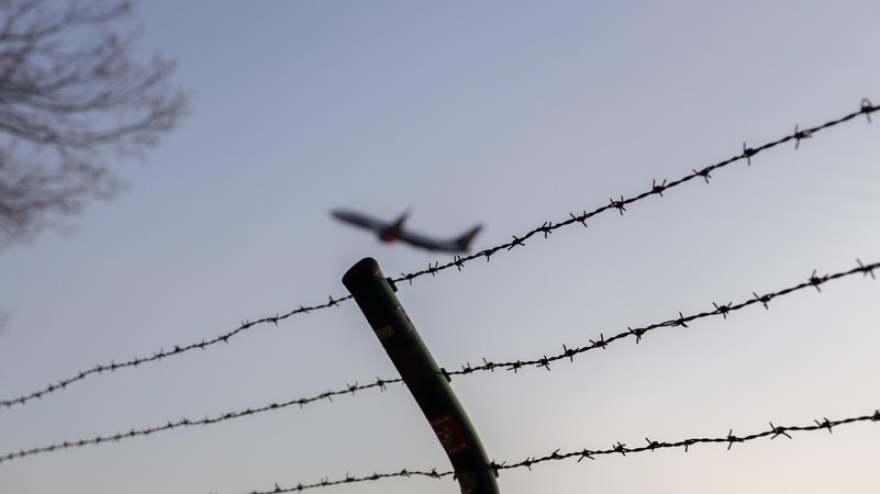 Ein Flugzeug fliegt in der Luft. Vordergründig sieht man einen Stacheldrahtzaun. | Bild: picture alliance / Bonn.digital | Marc John Ein Flugzeug fliegt in der Luft. Vordergründig sieht man einen Stacheldrahtzaun.