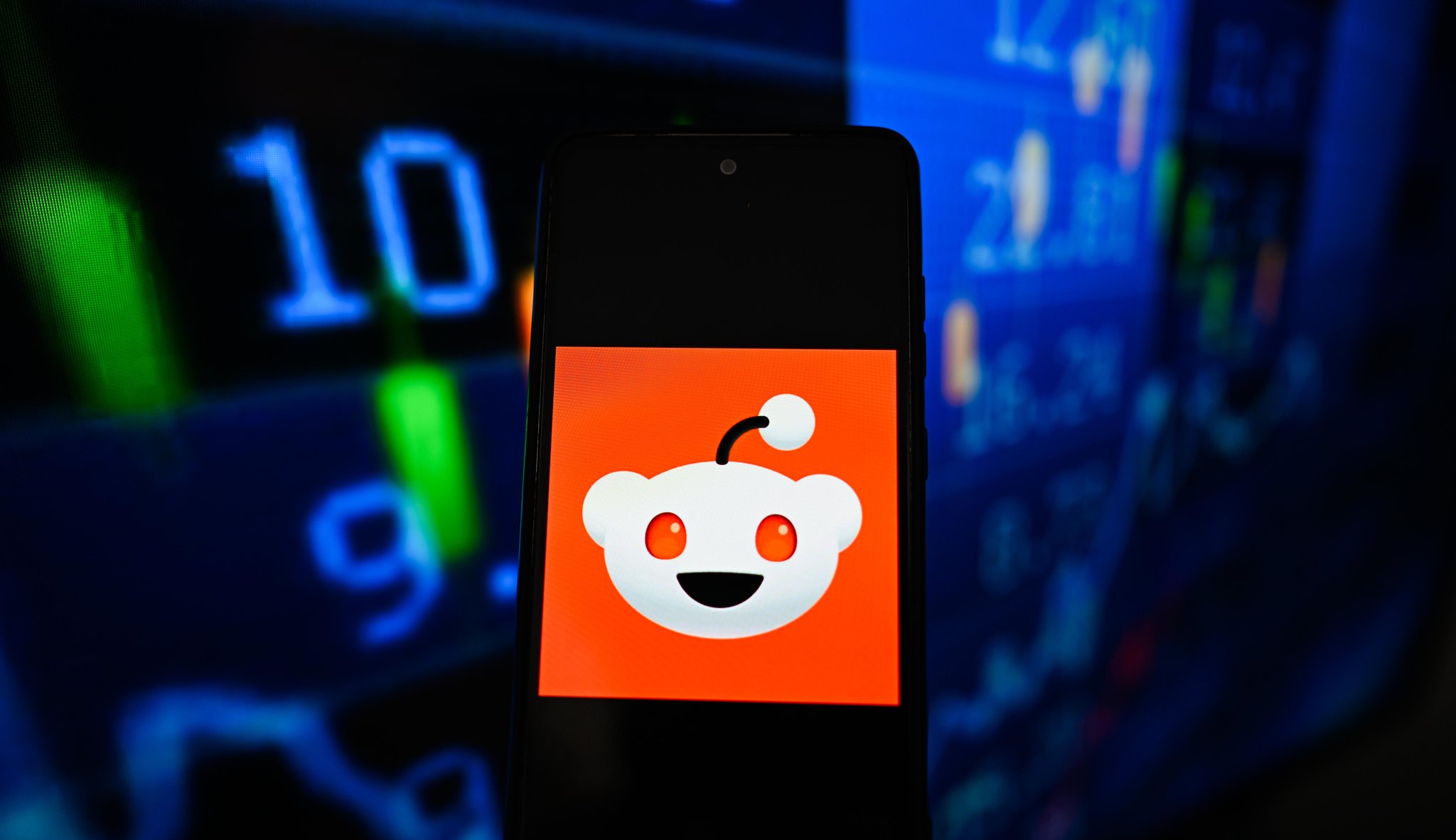 (Symbolbild) Auf dieser Fotoillustration ist das Reddit-Logo auf einem Smartphone zu sehen, im Hintergrund sind Börsenkurse abgebildet.