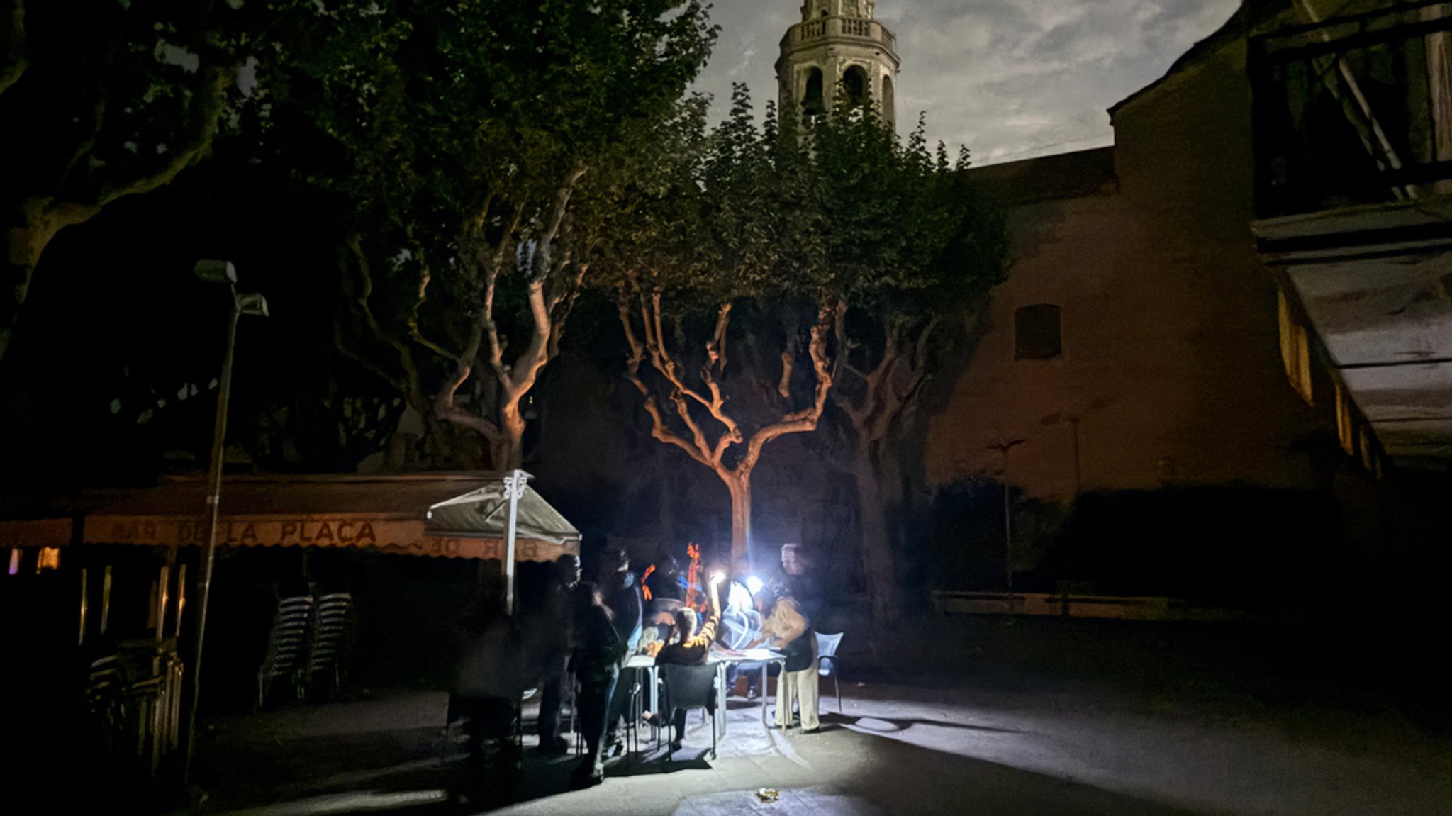 28.04.2025, Spanien, Premià de Mar: Auf der Placa de l'Ajuntament sitzen die Gäste eines Straßencafés in der Kleinstadt Premià de Mar bei Barcelona an der Kirche "San Cristòfol" bei einem Stromausfall im Licht von Taschenlampen und trinken lauwarmes Bier.