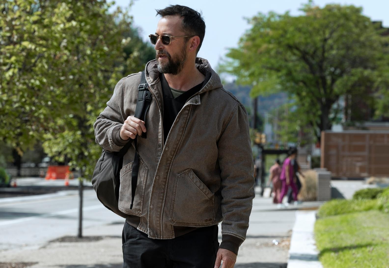 Noah Wyle alias läuft eine sonnige Straße entlang, den Rucksack locker über der rechten Schulter, Szene aus "The Pitt"