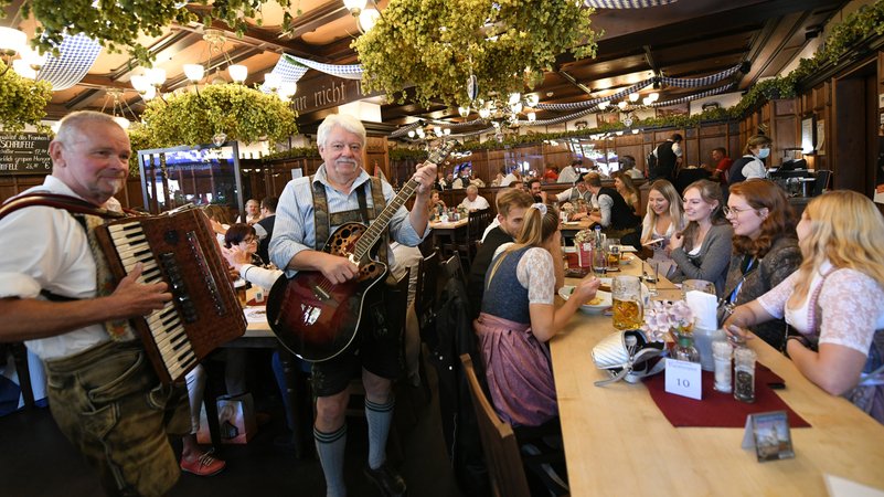 Gäste feiern an voll besetzten Tischen in einer Gaststätte; in der Mitte stehen zwei Musikanten in Tracht und spielen auf dem Akkordeon und der Gitarre. 