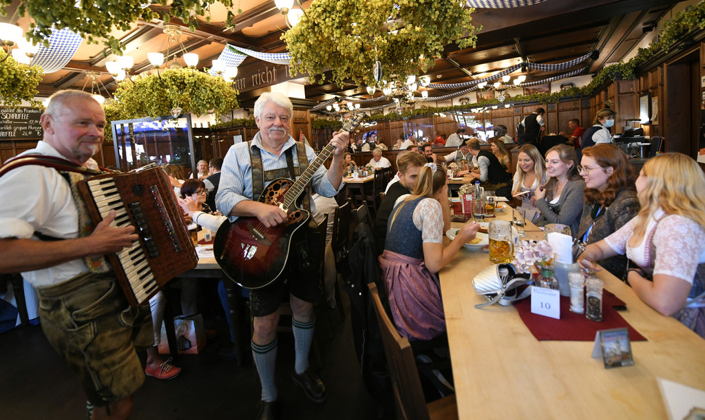Gäste feiern an voll besetzten Tischen in einer Gaststätte; in der Mitte stehen zwei Musikanten in Tracht und spielen auf dem Akkordeon und der Gitarre. 