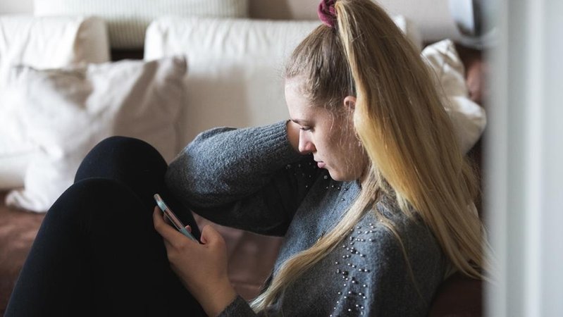 Eine junge Frau sitzt alleine in ihrer Wohnung und hält über ihr Smartphone Kontakt zu ihren Freunden und ihrer Familie.