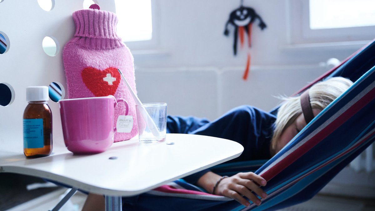 Ein Kind schläft in einer Hängematte. Auf einem Tisch stehen eine Wärmflasche, Hustensaft, Fieberthermometer und eine Tasse Tee. 