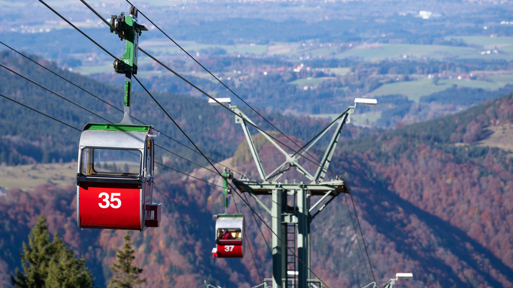 Die Kampenwandbahn aus einer Gondel gesehen: Vor und hinter einem Stützpfeiler hängt jeweils eine rote Gondel aus den 1950er-Jahren. Im Hintergrund Ausblick ins Chiemgau.