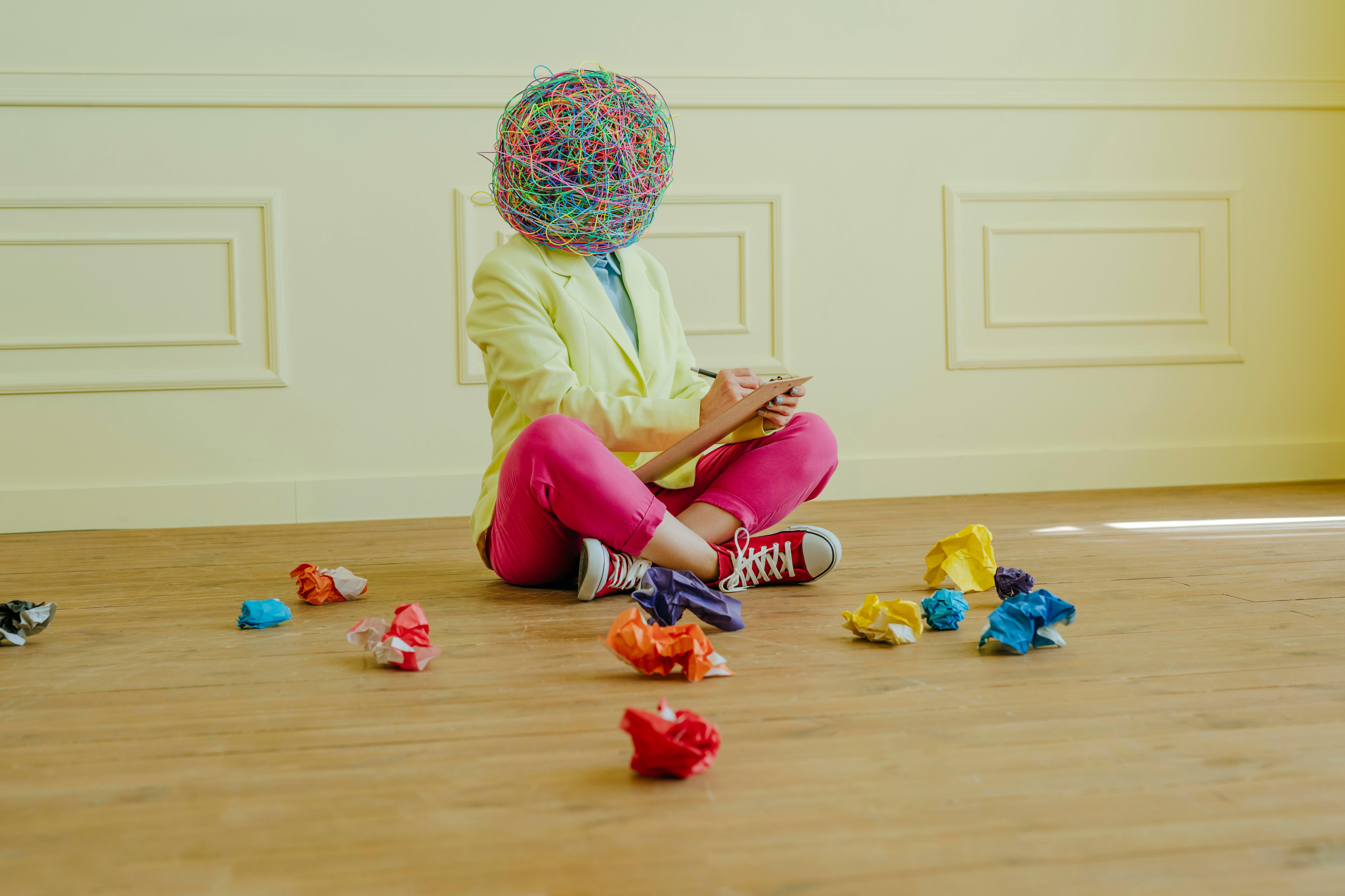 Frau mit einer Maske, die "chaotische Gedanken" zeigt, sitzt auf dem Boden zwischen zerknülltenm Papier, schreibt etwas auf.