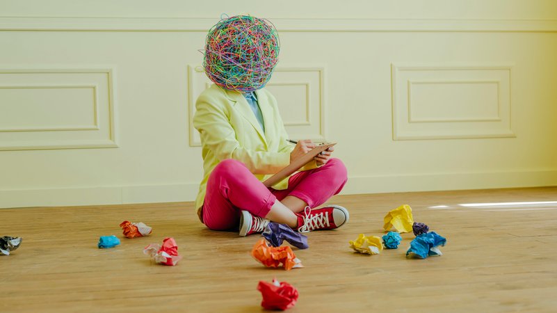 Frau mit einer Maske, die "chaotische Gedanken" zeigt, sitzt auf dem Boden zwischen zerknülltenm Papier, schreibt etwas auf. | Bild: picture alliance / Westend61 | Elisatim Frau mit einer Maske, die "chaotische Gedanken" zeigt, sitzt auf dem Boden zwischen zerknülltenm Papier, schreibt etwas auf.