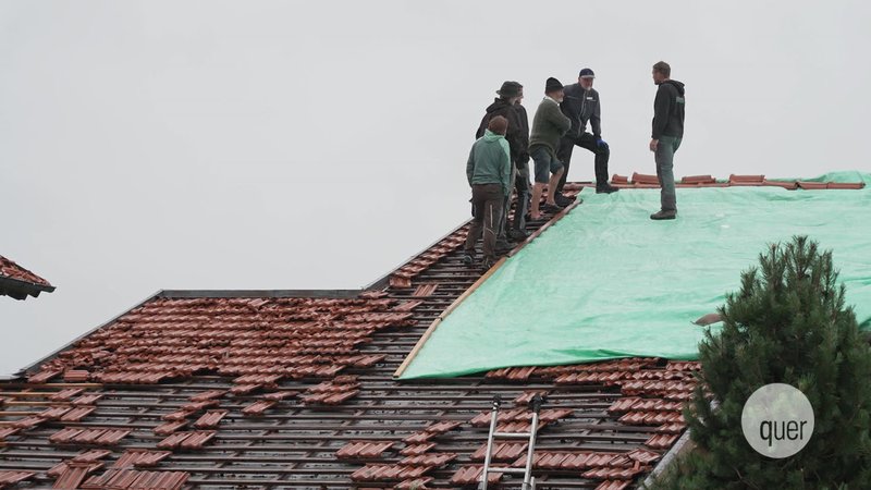 Menschen stehen auf einem Dach und decken ein Loch mit einer Plane ab.