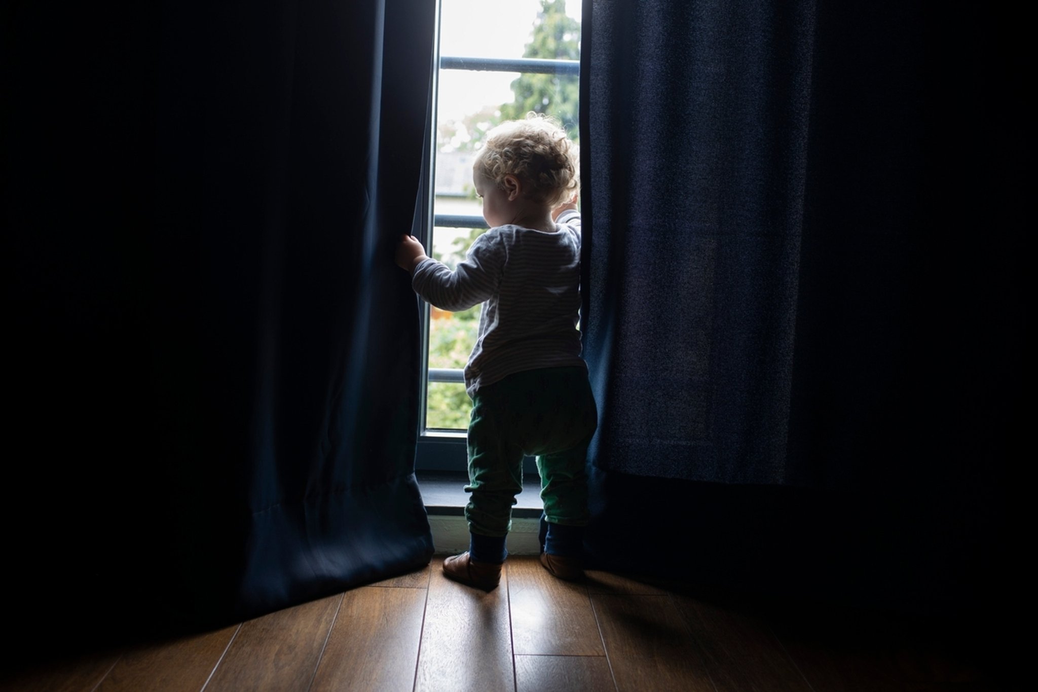 Symbolbild: Kleines Kind in einem dunkel Zimmer schaut durch die Vorhänge hindurch aus dem Fenster