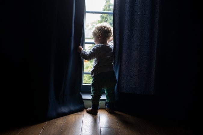Symbolbild: Kleines Kind in einem dunkel Zimmer schaut durch die Vorhänge hindurch aus dem Fenster