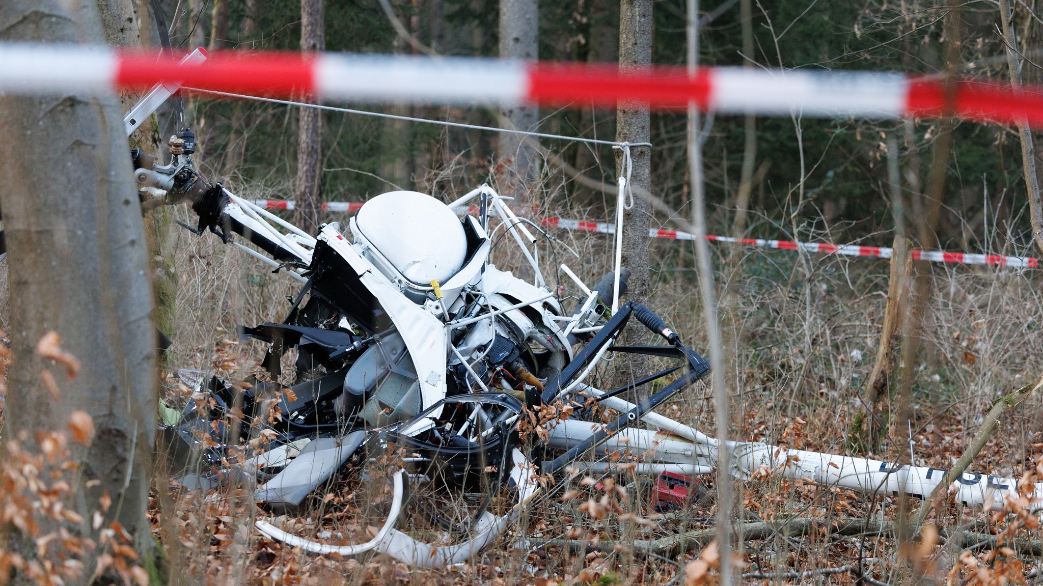 Wrackteile eines Hubschraubers liegen im Wald, Rot-Weißes-Absperrband ist an Bäumen angebracht