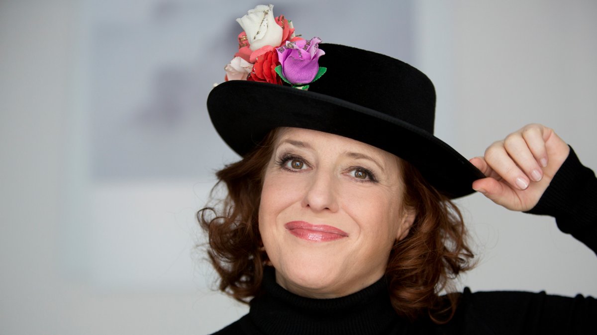 Luise Kinseher mit schwarzem Hut und Blüte lächelt keck. 