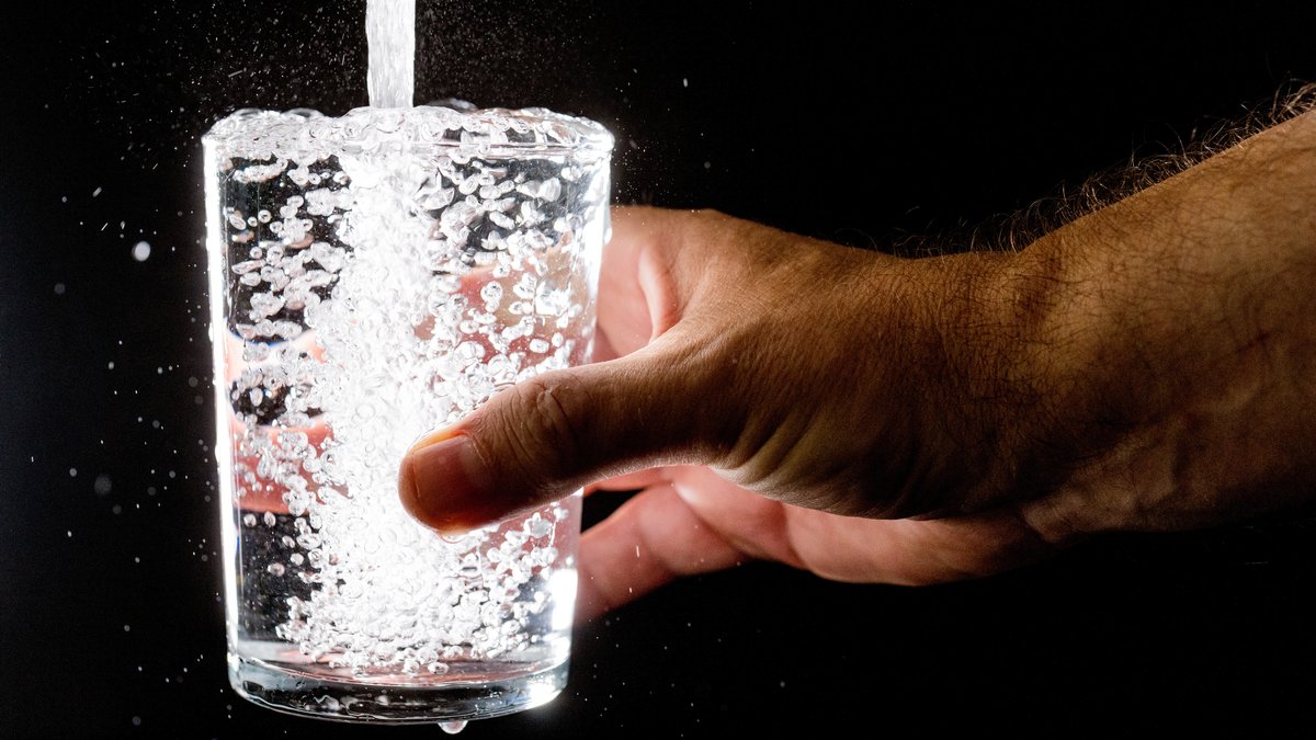 Erleuchtetes Wasserglas, das mit Wasser aus dem Hahn gefüllt wird, gehalten von Hand; Kritische Infrastrukturen sind nicht nur Strom und Energie, auch Wasser gehört dazu.