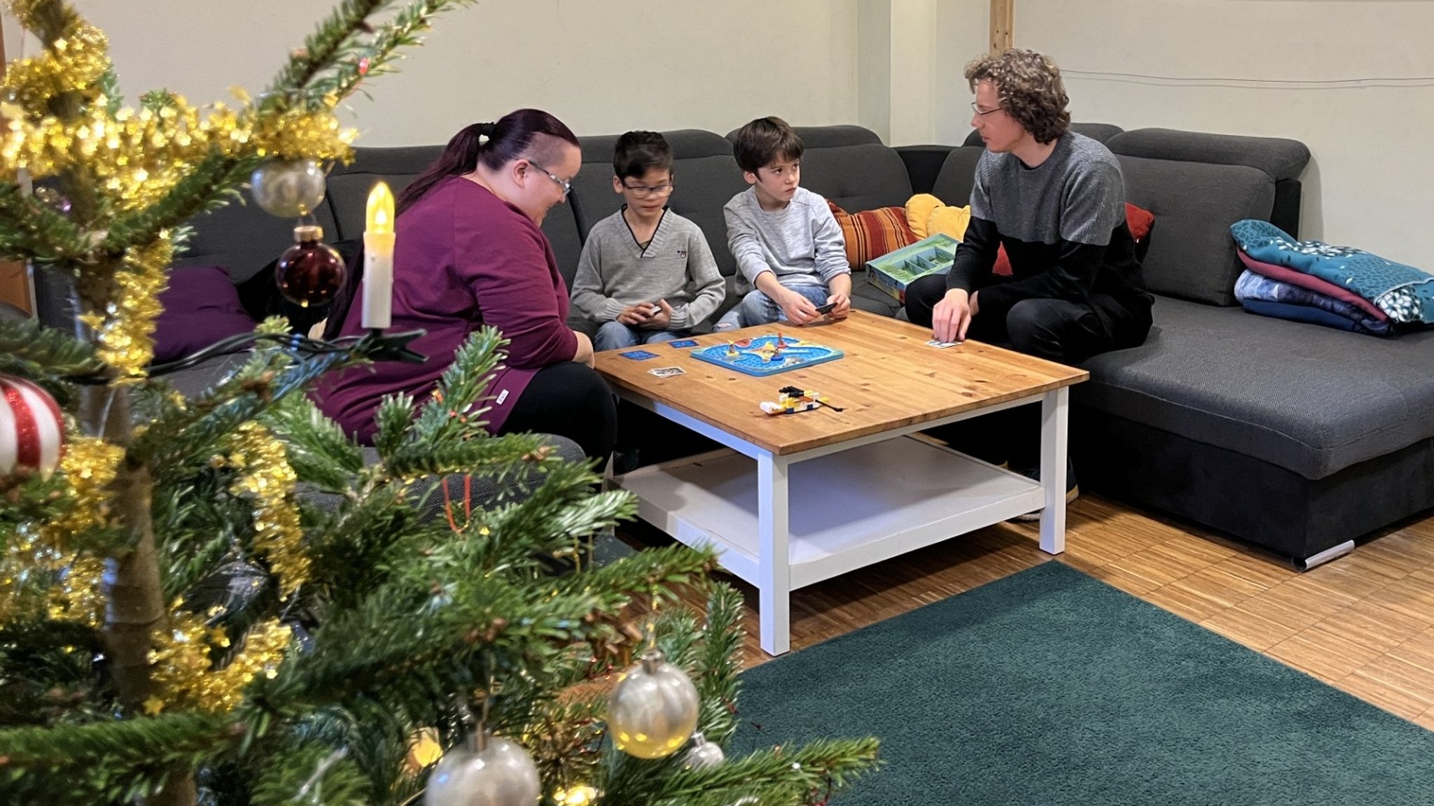 Zwei Kinder und zwei Erwachsene sitzen um einen Couchtisch, im Vordergrund ein Weihnachtsbaum.