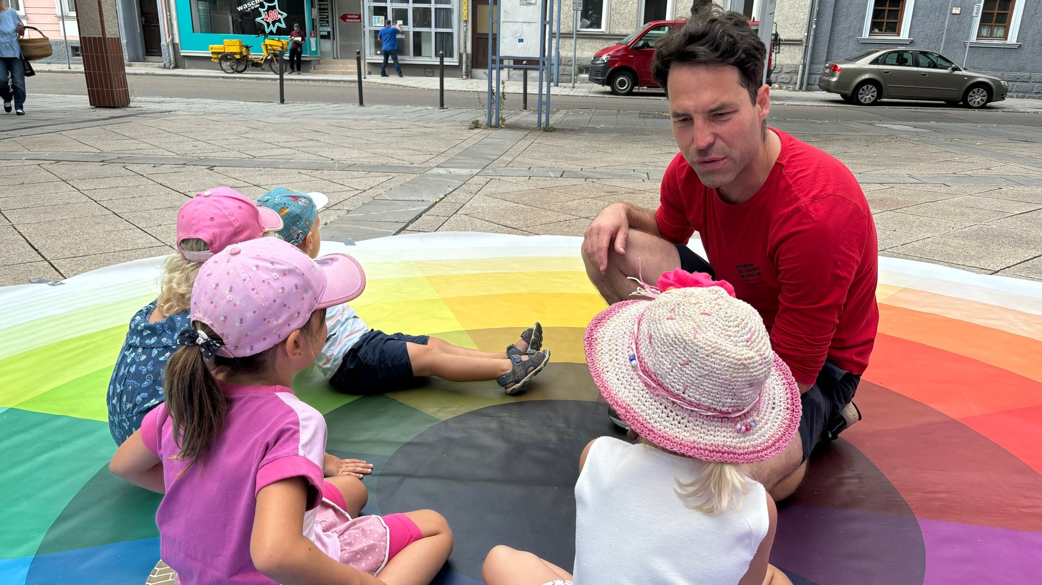 Kunstpädagoge Philipp Schneider sitzt mit vier Kindern auf einem Kreis aus verschiedenen Farben.